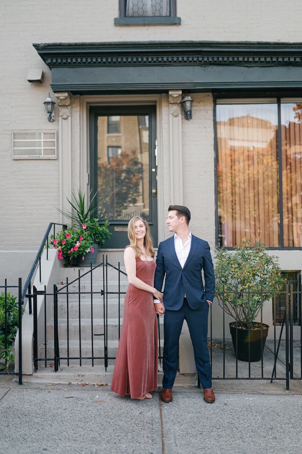 Eli and Georgia, in front of a Brooklyn brownstone, holding hands. Eli is looking at Georgia, and Georgia is looking at the camera.